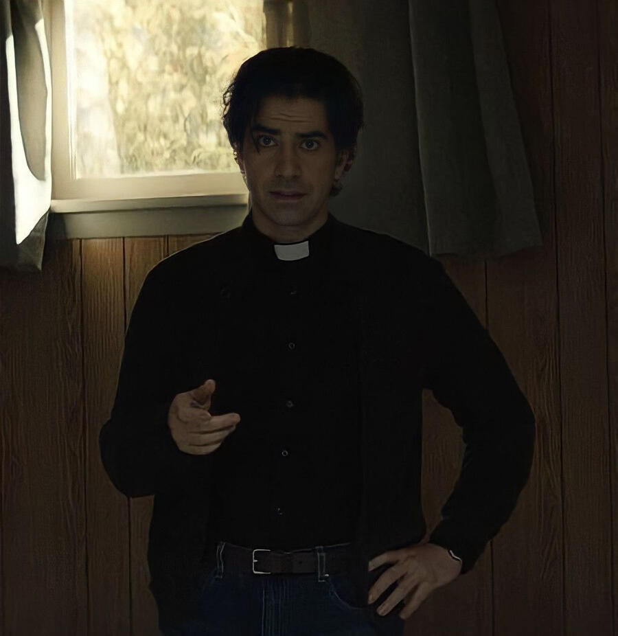 screenshot of paul hill from midnight mass. he's wearing a black clergy shirt a black cardigan and blue jeans. he has short wavy black hair that cascades into his face and he appears to be wearing some eyeliner. he has thick eyebrows and puppy dog eyes