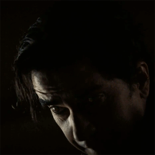 gif from midnight mass of paul in a dark room the lighting reflects in his eyes as if they were cat eyes. his eyes are glowing like little spheres. he moves his head around while he speaks his hair spilling into his eyes